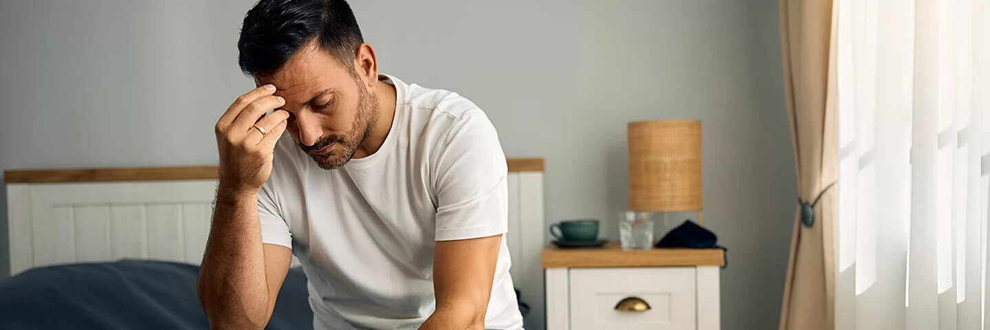 Ein junger Mann sitzt auf seinem Bett im Schlafzimmer. Mit gesenktem Kopf stützt er den rechten Ellenbogen auf dem rechten Knie ab und fasst sich mit der rechten Hand an die Stirn.