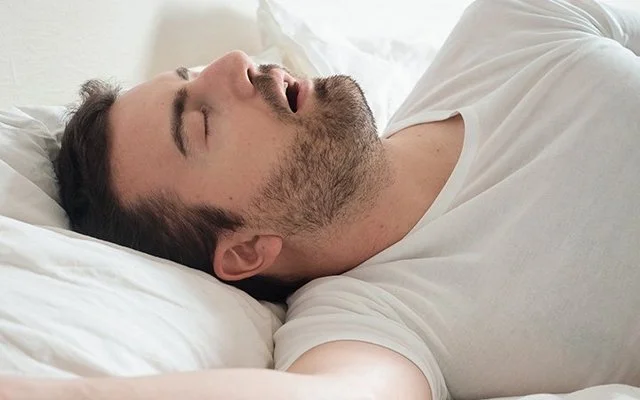 Ein Mann liegt mit offenem Mund im Bett und schnarcht.