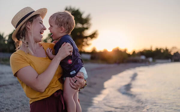 Mutter ist mit ihrem Baby im Urlaub am Strand, beide lachen sich fröhlich an.