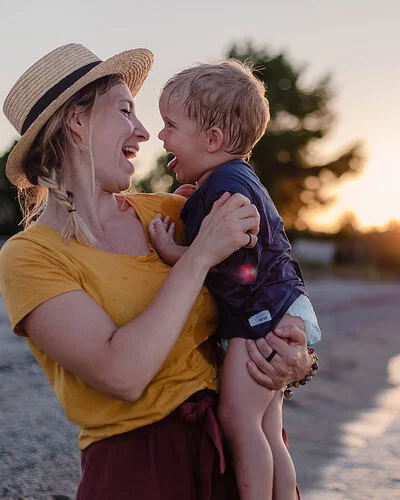 Mutter ist mit ihrem Baby im Urlaub am Strand, beide lachen sich fröhlich an.