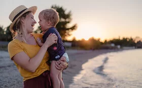 Mutter ist mit ihrem Baby im Urlaub am Strand, beide lachen sich fröhlich an.