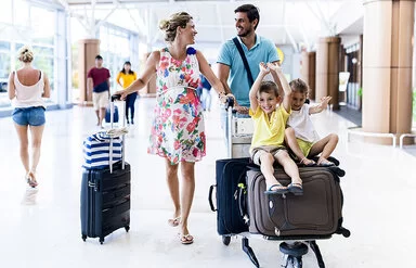 Das Bild zeigt eine glückliche Familie mit zwei Kindern am Flughafen. Die schwangere Mutter und der Vater schieben einen Gepäckwagen, auf dem ihre Koffer sowie die Kinder sitzen. Diese lachen und strecken fröhlich die Arme in die Luft.