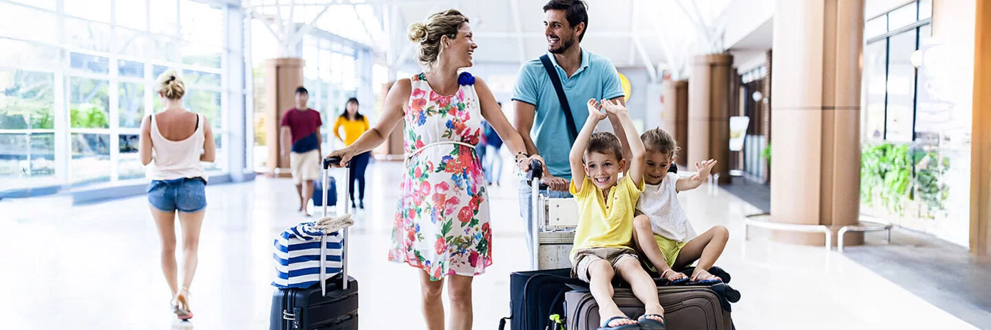 Das Bild zeigt eine glückliche Familie mit zwei Kindern am Flughafen. Die schwangere Mutter und der Vater schieben einen Gepäckwagen, auf dem ihre Koffer sowie die Kinder sitzen. Diese lachen und strecken fröhlich die Arme in die Luft.