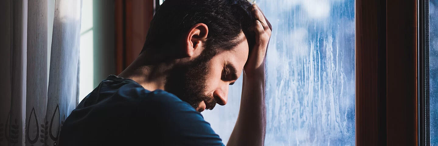 Ein Mann mit Depressionen steht vor einem Fenster und hält sich den Kopf.