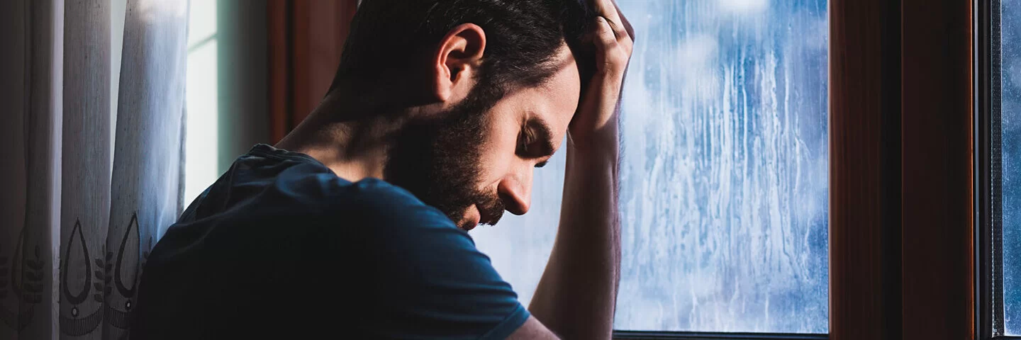 Ein Mann mit Depressionen steht vor einem Fenster und hält sich den Kopf.