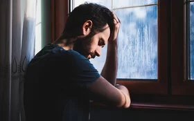 Ein Mann mit Depressionen steht vor einem Fenster und hält sich den Kopf.