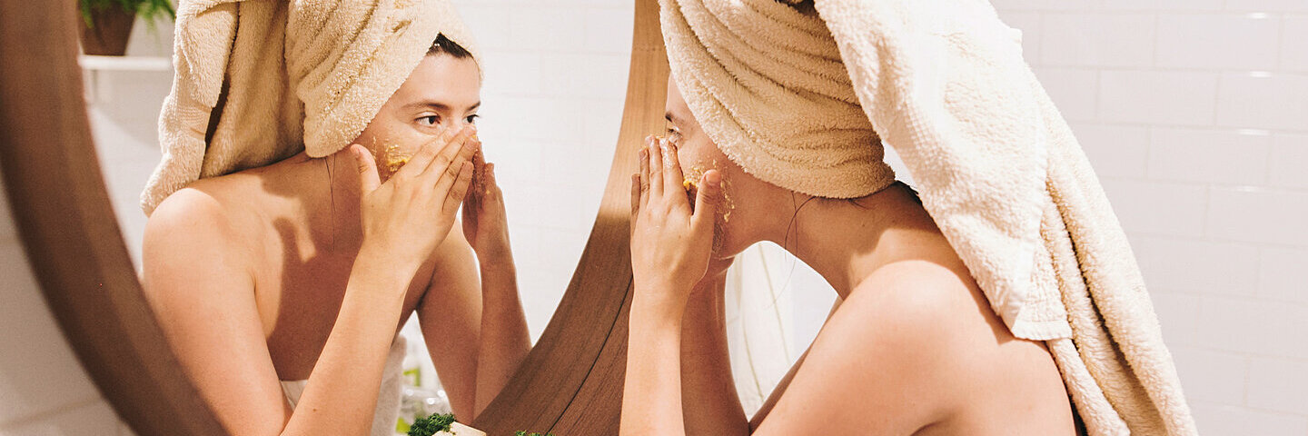 Eine in Handtüchern gekleidete Frau steht im Bad vorm Spiegel und benutzt ein Naturkosmetik-Peeling.