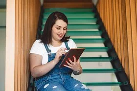 Eine junge Frau sitzt auf einer Treppe und sucht am Tablet nach Online-Selbsthilfe.
