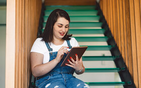 Eine junge Frau sitzt auf einer Treppe und sucht am Tablet nach Online-Selbsthilfe.