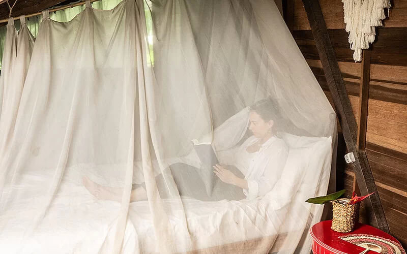 Eine Frau liegt in einem Holzhaus auf dem Bett und liest. Das Bett ist von einem Moskitonetz umgeben.