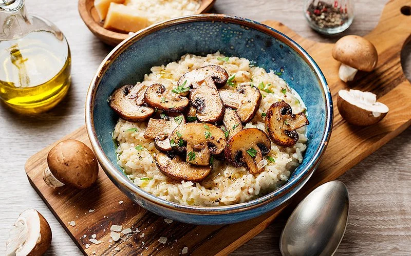 Auf einem Tisch steht eine Schüssel mit Risotto. Gebratene braune Champignons und Petersilie dienen als Topping.