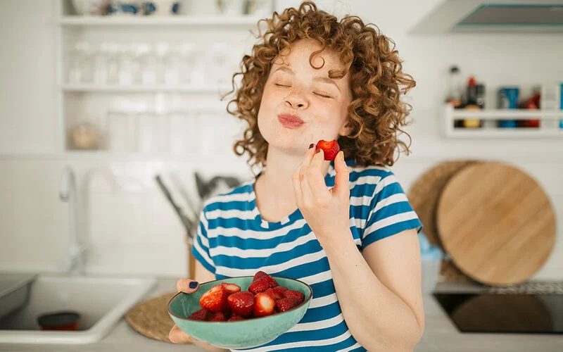Eine Frau steht in der Küche und isst genüßlich Erdbeeren aus einer Schüssel.