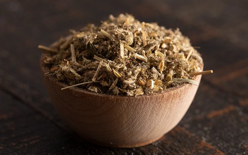 Auf einer Holztischplatte steht eine Holzschale. Diese ist mit gehackten und getrockneten Andorn-Stängeln, -Blättern und -Blüten gefüllt.