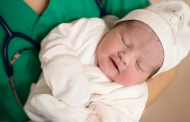 Eine medizinische Fachkraft in grüner Kleidung hält ein schlafendes Neugeborenes im Arm. Das Baby trägt weiße Kleidung, eine Mütze und Handschuhe.