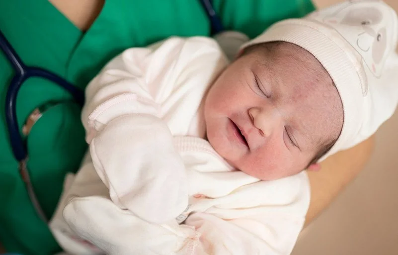 Eine medizinische Fachkraft in grüner Kleidung hält ein schlafendes Neugeborenes im Arm. Das Baby trägt weiße Kleidung, eine Mütze und Handschuhe.