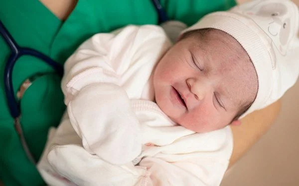 Eine medizinische Fachkraft in grüner Kleidung hält ein schlafendes Neugeborenes im Arm. Das Baby trägt weiße Kleidung, eine Mütze und Handschuhe.