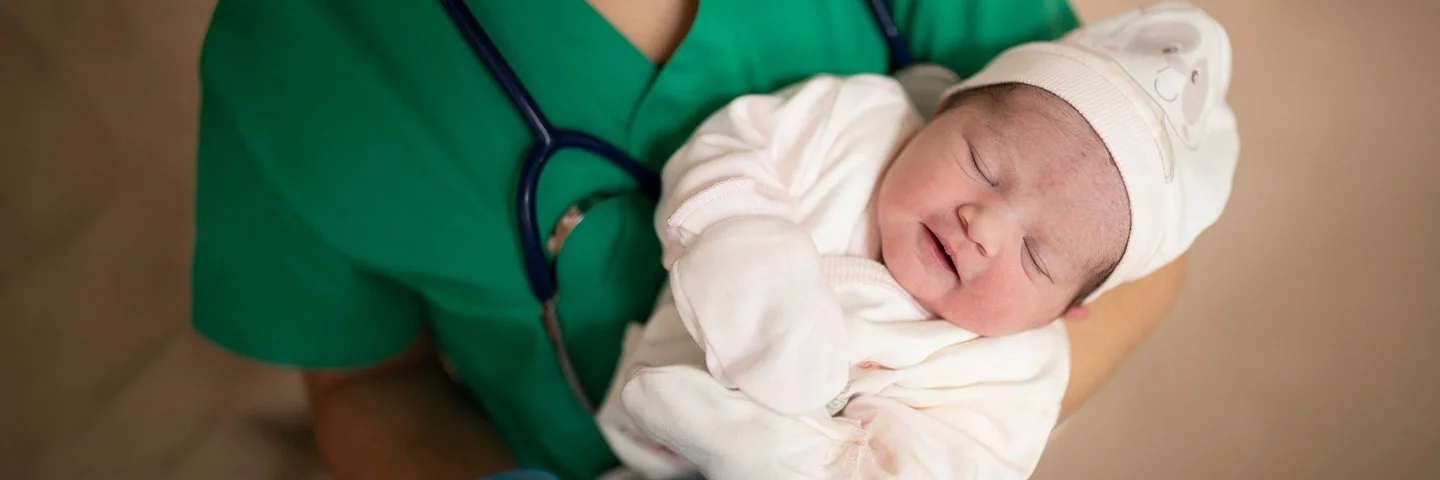 Eine medizinische Fachkraft in grüner Kleidung hält ein schlafendes Neugeborenes im Arm. Das Baby trägt weiße Kleidung, eine Mütze und Handschuhe.