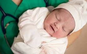 Eine medizinische Fachkraft in grüner Kleidung hält ein schlafendes Neugeborenes im Arm. Das Baby trägt weiße Kleidung, eine Mütze und Handschuhe.