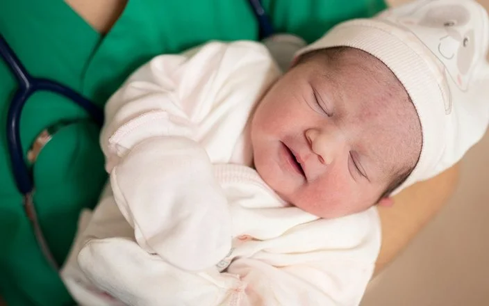 Eine medizinische Fachkraft in grüner Kleidung hält ein schlafendes Neugeborenes im Arm. Das Baby trägt weiße Kleidung, eine Mütze und Handschuhe.