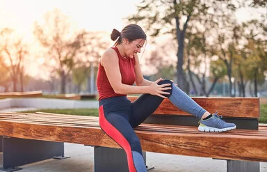 Eine junge Sportlerin sitzt auf einer Parkbank und hält unter Schmerzen das linke Knie in beiden Händen.