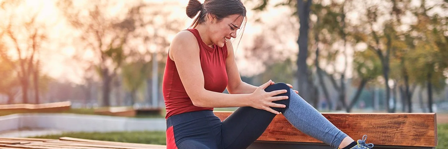 Eine junge Sportlerin sitzt auf einer Parkbank und hält unter Schmerzen das linke Knie in beiden Händen.