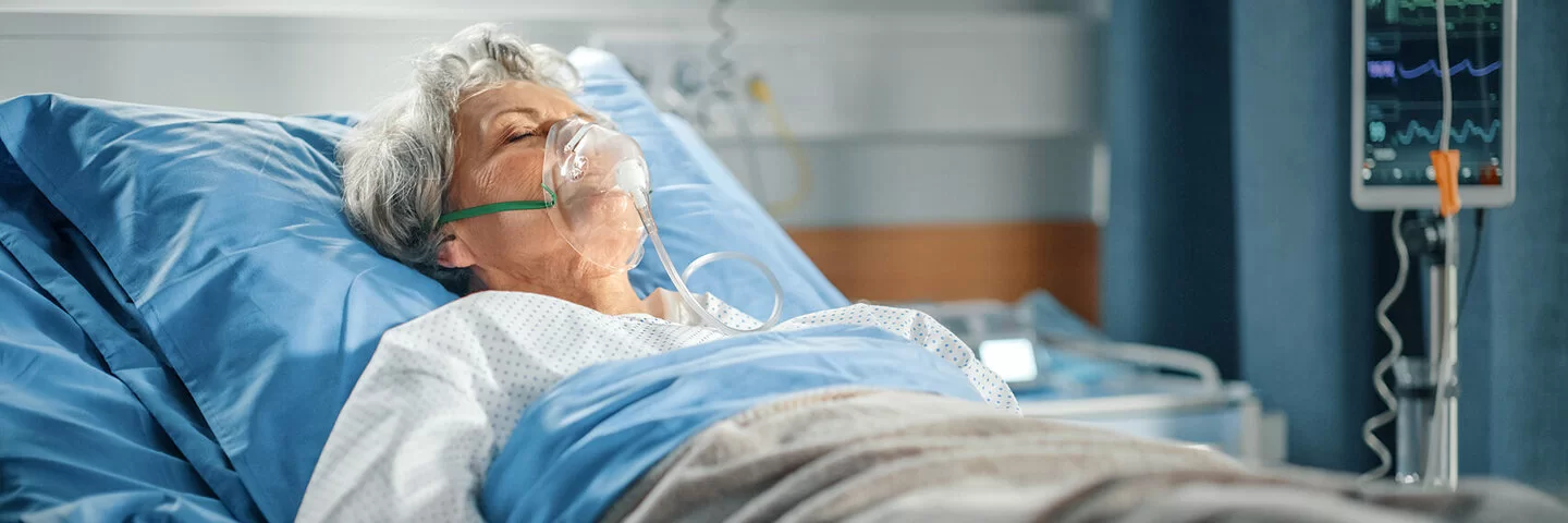 Eine Frau, die an einer Lungenembolie erkrankt ist, liegt mit Beatmungsmaske in einem Krankenhausbett.