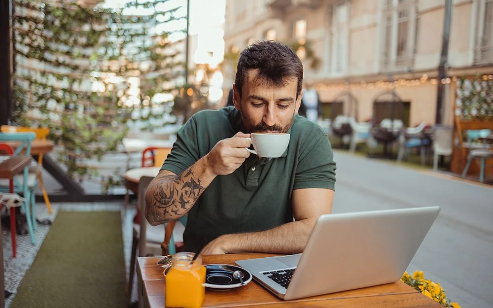 In einem Straßencafé sitzt ein Mann an einem Tisch, vor sich einen aufgeklappten Laptop, und trinkt eine Tasse Kaffee.