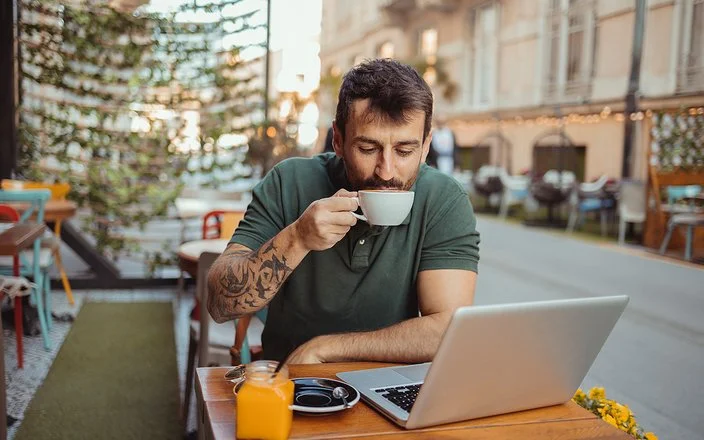 In einem Straßencafé sitzt ein Mann an einem Tisch, vor sich einen aufgeklappten Laptop, und trinkt eine Tasse Kaffee.
