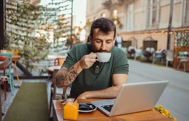 In einem Straßencafé sitzt ein Mann an einem Tisch, vor sich einen aufgeklappten Laptop, und trinkt eine Tasse Kaffee.