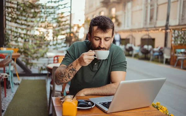 In einem Straßencafé sitzt ein Mann an einem Tisch, vor sich einen aufgeklappten Laptop, und trinkt eine Tasse Kaffee.