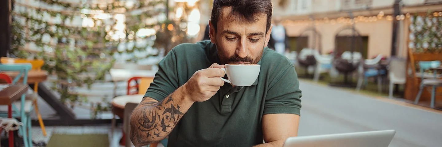 In einem Straßencafé sitzt ein Mann an einem Tisch, vor sich einen aufgeklappten Laptop, und trinkt eine Tasse Kaffee.