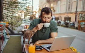 In einem Straßencafé sitzt ein Mann an einem Tisch, vor sich einen aufgeklappten Laptop, und trinkt eine Tasse Kaffee.