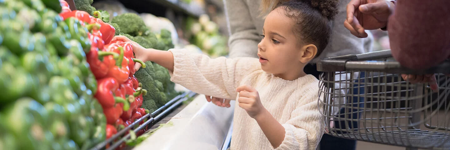 Kleines Mädchen darf beim kauf von Gemüse helfen und greift zur roten Paprika.