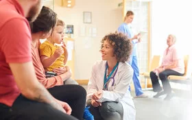 Eine Kinderärztin begrüßt ein Kind mit Borkenflechte und dessen Eltern im Flur.