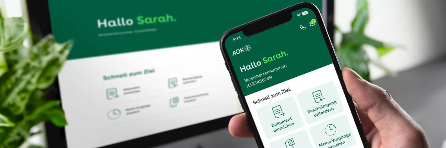 Eine Person hält ein Smartphone in der Hand. Auf dem Bildschirm ist die geöffnete App „Meine AOK“ zu sehen. Im Hintergrund steht ein PC-Monitor, auf dem ebenfalls das Online-Portal „Meine AOK“ zu sehen ist.