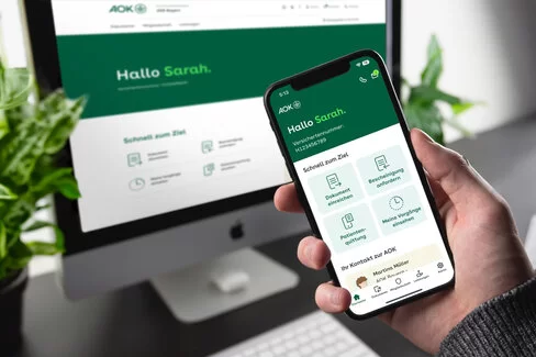 Eine Person hält ein Smartphone in der Hand. Auf dem Bildschirm ist die geöffnete App „Meine AOK“ zu sehen. Im Hintergrund steht ein PC-Monitor, auf dem ebenfalls das Online-Portal „Meine AOK“ zu sehen ist.