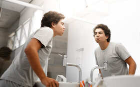 Ein junger Mann benutzt eine Mundspülung und schaut dabei in den Spiegel im Bad.