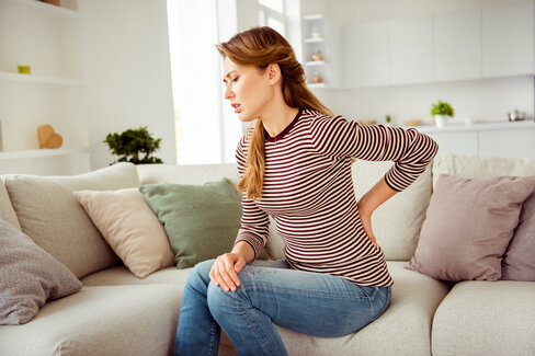 Eine Frau sitzt auf dem Sofa und hält sich den unteren Rücken. Sie leidet an Rückenschmerzen.