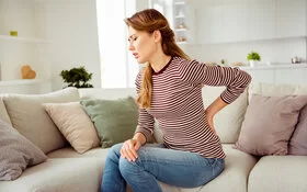 Eine Frau sitzt auf dem Sofa und hält sich den unteren Rücken. Sie leidet an Rückenschmerzen.