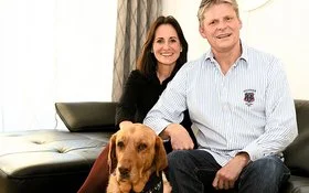 Jörg Zuber sitzt mit seiner Frau Tanja Eggers und seinem Labrador Ginny auf einem schwarzen Ledersofa im Wohnzimmer.