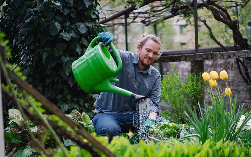 Ein junger Mann hockt in einem Garten und gießt Kräuter mit einer grünen Gießkanne.