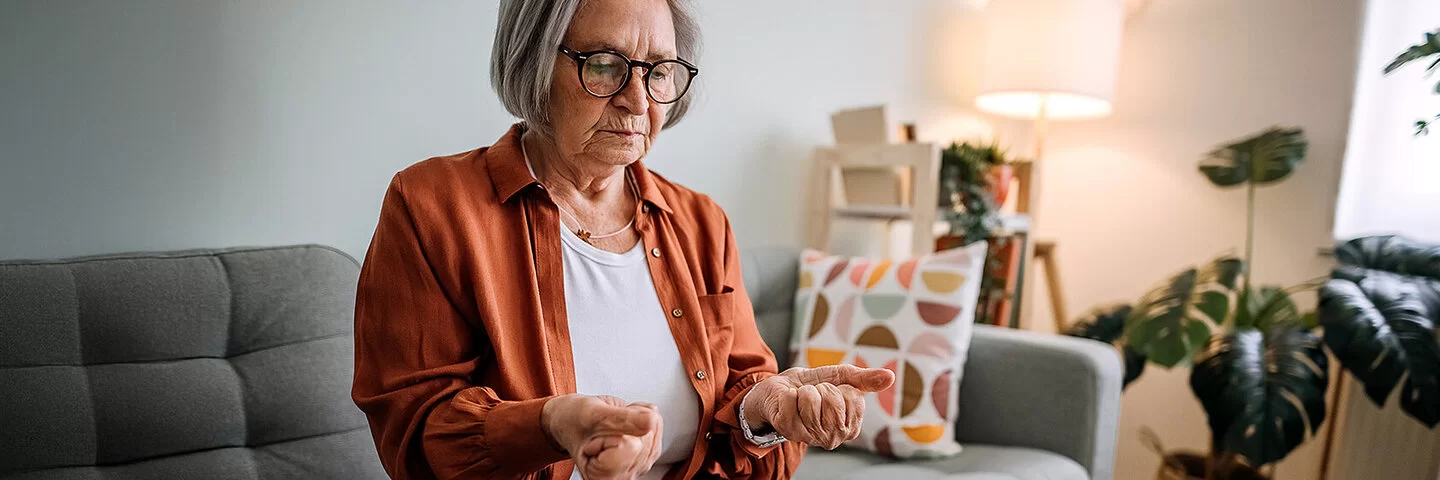 Ältere Frau mit Arthrose sitzt auf einem Sofa in ihrem Zuhause.Sie betrachtet ihre zu Fäusten zusammengeballten Hände, die sie von sich gestreckt hat.