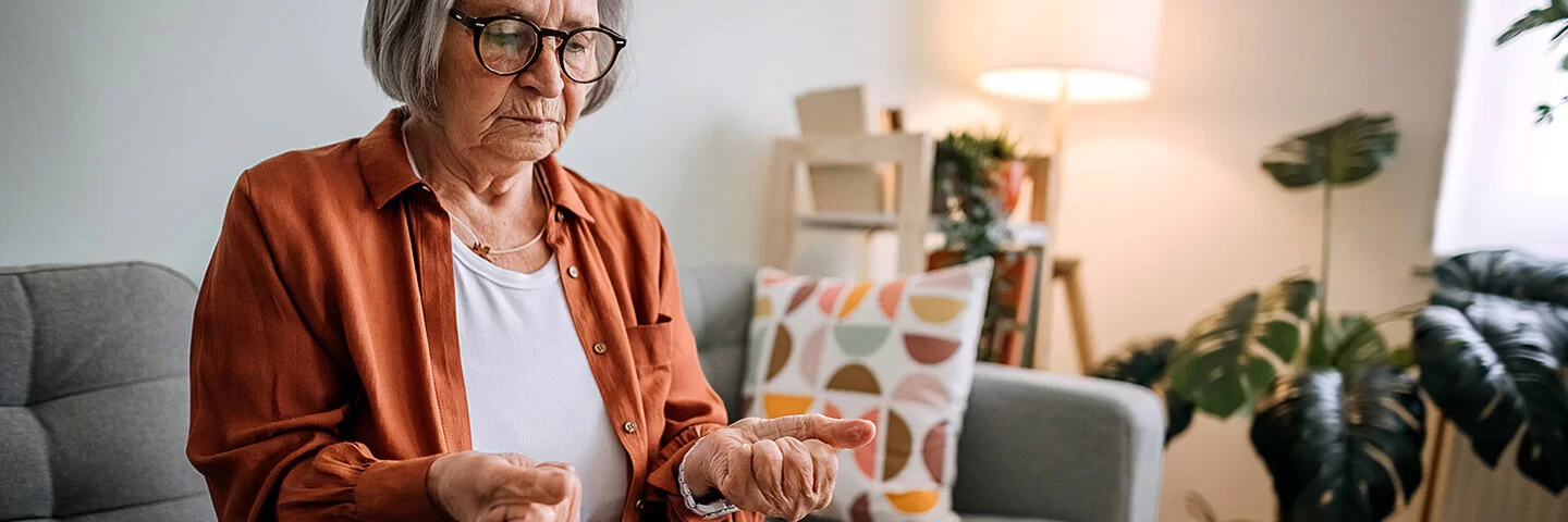 Ältere Frau mit Arthrose sitzt auf einem Sofa in ihrem Zuhause.Sie betrachtet ihre zu Fäusten zusammengeballten Hände, die sie von sich gestreckt hat.