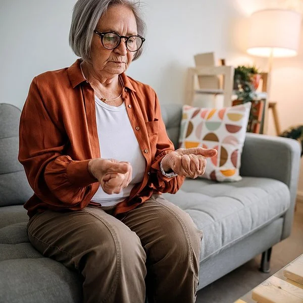 Ältere Frau mit Arthrose sitzt auf einem Sofa in ihrem Zuhause.Sie betrachtet ihre zu Fäusten zusammengeballten Hände, die sie von sich gestreckt hat.
