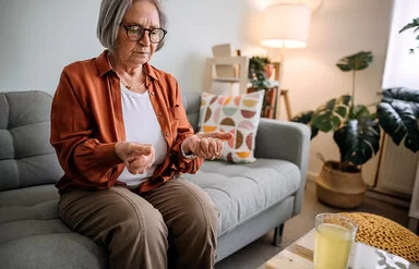 Ältere Frau mit Arthrose sitzt auf einem Sofa in ihrem Zuhause.Sie betrachtet ihre zu Fäusten zusammengeballten Hände, die sie von sich gestreckt hat.