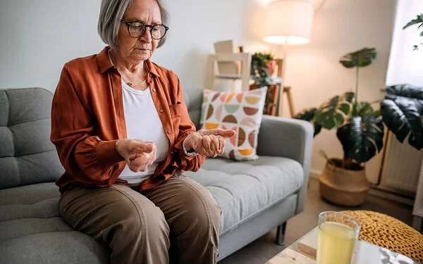 Ältere Frau mit Arthrose sitzt auf einem Sofa in ihrem Zuhause.Sie betrachtet ihre zu Fäusten zusammengeballten Hände, die sie von sich gestreckt hat.