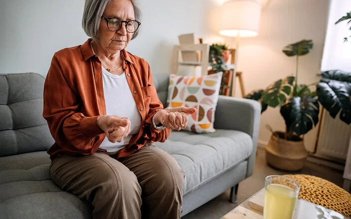Ältere Frau mit Arthrose sitzt auf einem Sofa in ihrem Zuhause.Sie betrachtet ihre zu Fäusten zusammengeballten Hände, die sie von sich gestreckt hat.