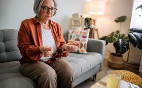 Ältere Frau mit Arthrose sitzt auf einem Sofa in ihrem Zuhause.Sie betrachtet ihre zu Fäusten zusammengeballten Hände, die sie von sich gestreckt hat.