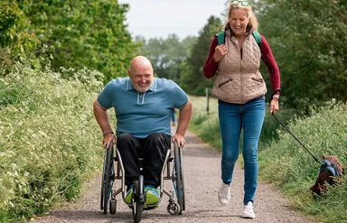 Ein Mann im Rollstuhl und eine Frau mit Hund sind gemeinsam auf einem Naturweg unterwegs.