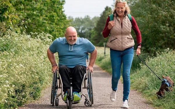 Ein Mann im Rollstuhl und eine Frau mit Hund sind gemeinsam auf einem Naturweg unterwegs. 
